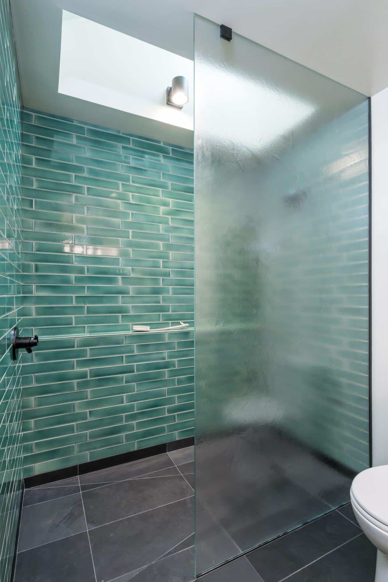Modern bathroom with green subway tile walls, frosted glass shower divider, black floor tiles, and a ceiling light.