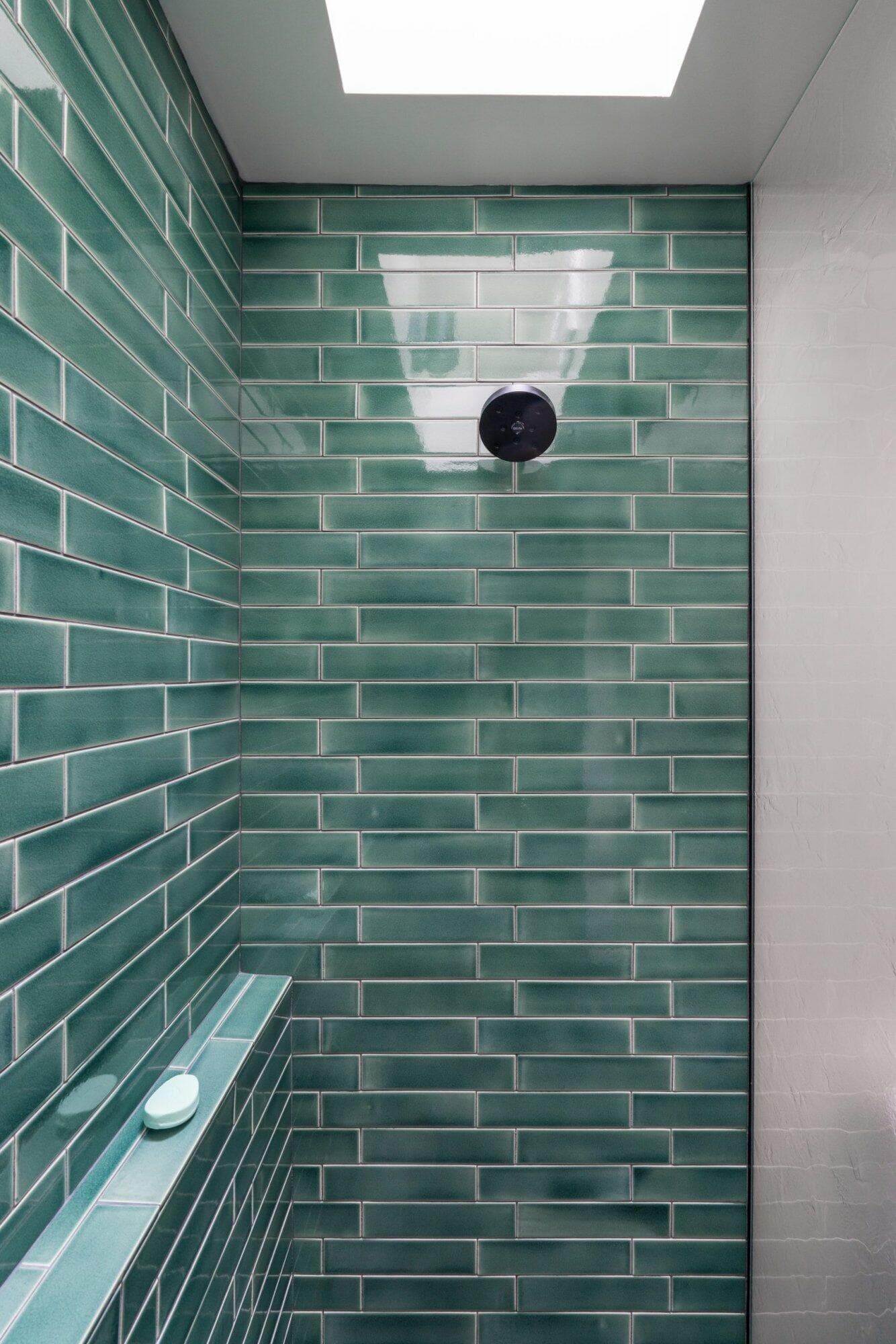 A shower area with green subway tiles, a black showerhead, and a small shelf holding a soap bar. There is a skylight above, allowing natural light to enter.