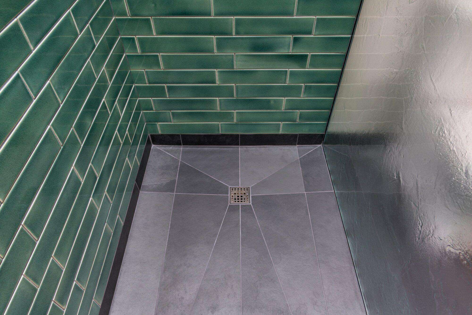 A tiled shower corner with green subway-style tiles on the wall and gray floor tiles. There is a square drain in the center of the floor.