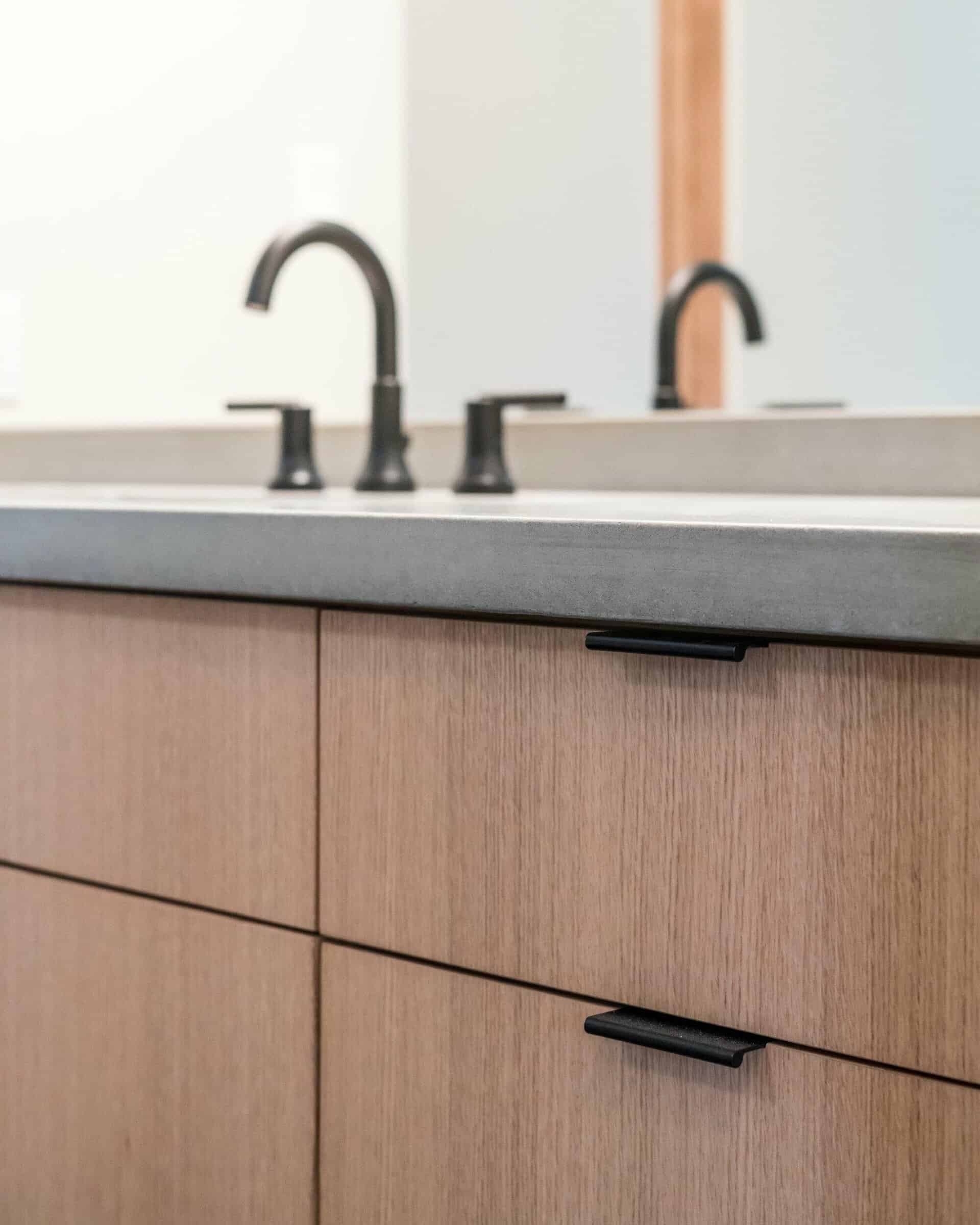 Close-up of a bathroom vanity with light wood cabinetry, black handles, and a gray countertop. A black faucet and a partial reflection in the mirror are visible above the counter.