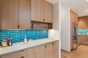 A modern kitchen with light wood cabinets, turquoise subway tile backsplash, and stainless steel appliances. Various bottles and glasses are displayed on the counter and shelves.