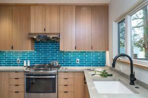 Modern kitchen with light wood cabinets, turquoise subway tile backsplash, stainless steel stove with hood, black faucet, and large window above a white sink. Countertop has vegetables and utensils.