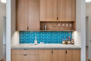 A home bar setup featuring wooden cabinets, blue tile backsplash, and a countertop with various bottles, glasses, and a shaker.