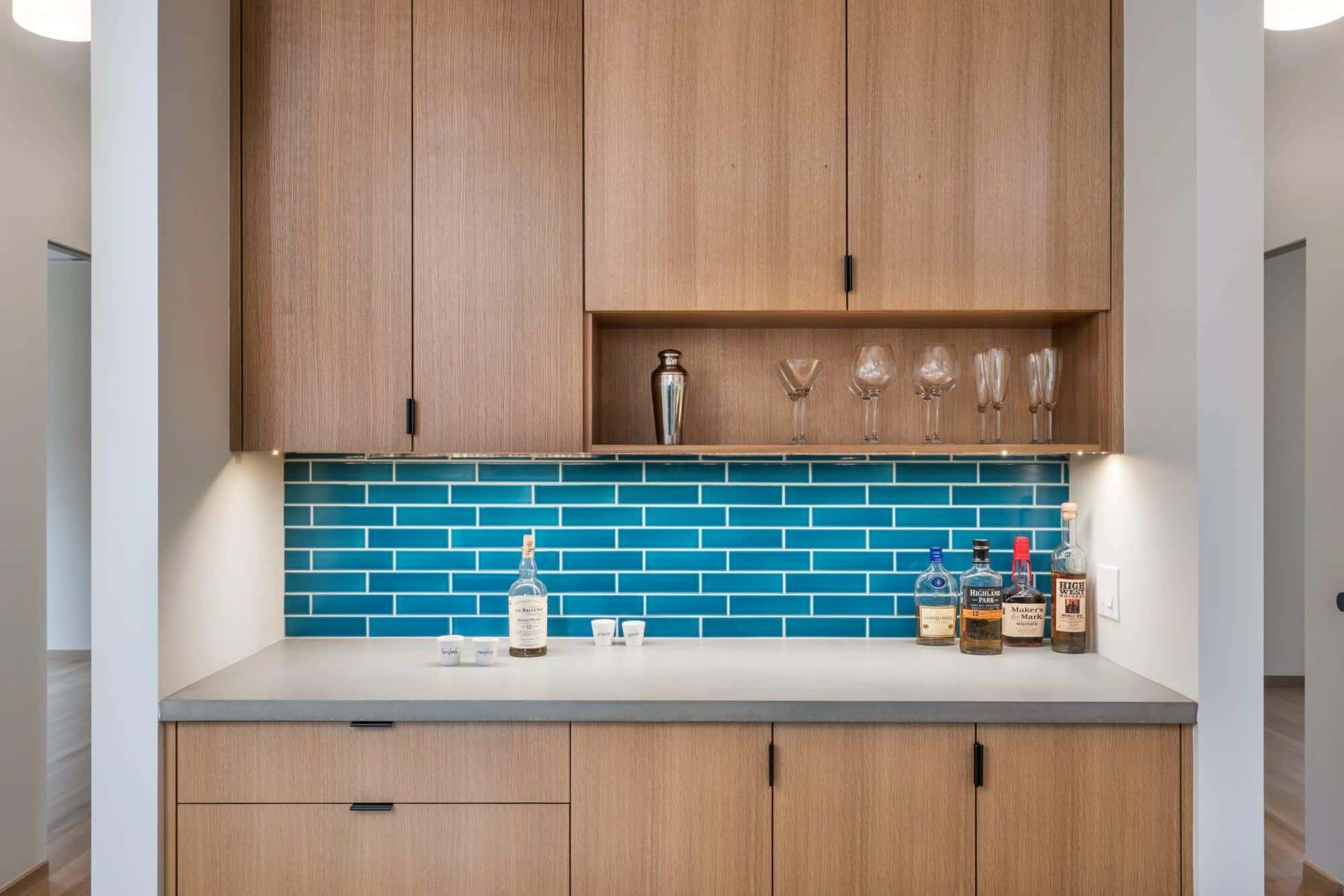 A home bar setup featuring wooden cabinets, blue tile backsplash, and a countertop with various bottles, glasses, and a shaker. A home bar setup featuring wooden cabinets, blue tile backsplash, and a countertop with various bottles, glasses, and a shaker.