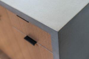 Close-up of a wooden cabinet corner with a smooth, light gray countertop. The cabinet has a black handle on the drawer.