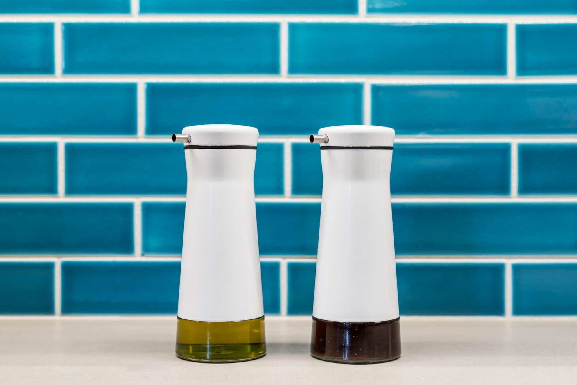 Two white ceramic dispensers with stainless steel nozzles, one filled with olive oil and the other with balsamic vinegar, placed on a countertop with a blue subway tile backsplash. Two white ceramic dispensers with stainless steel nozzles, one filled with olive oil and the other with balsamic vinegar, placed on a countertop with a blue subway tile backsplash.