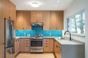 Modern kitchen with wooden cabinets, stainless steel appliances, blue tile backsplash, and a large window over the sink.