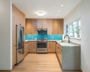 Modern kitchen with light wooden cabinets, stainless steel appliances, turquoise subway tile backsplash, and light hardwood floor. A window on the right side provides natural light.