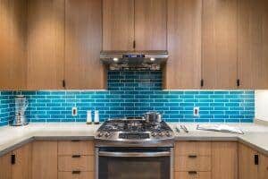 Modern kitchen with light wood cabinets, blue subway tile backsplash, stainless steel range hood, gas stove, and a countertop with a blender, salt and pepper shakers, a pot, and an open cookbook.