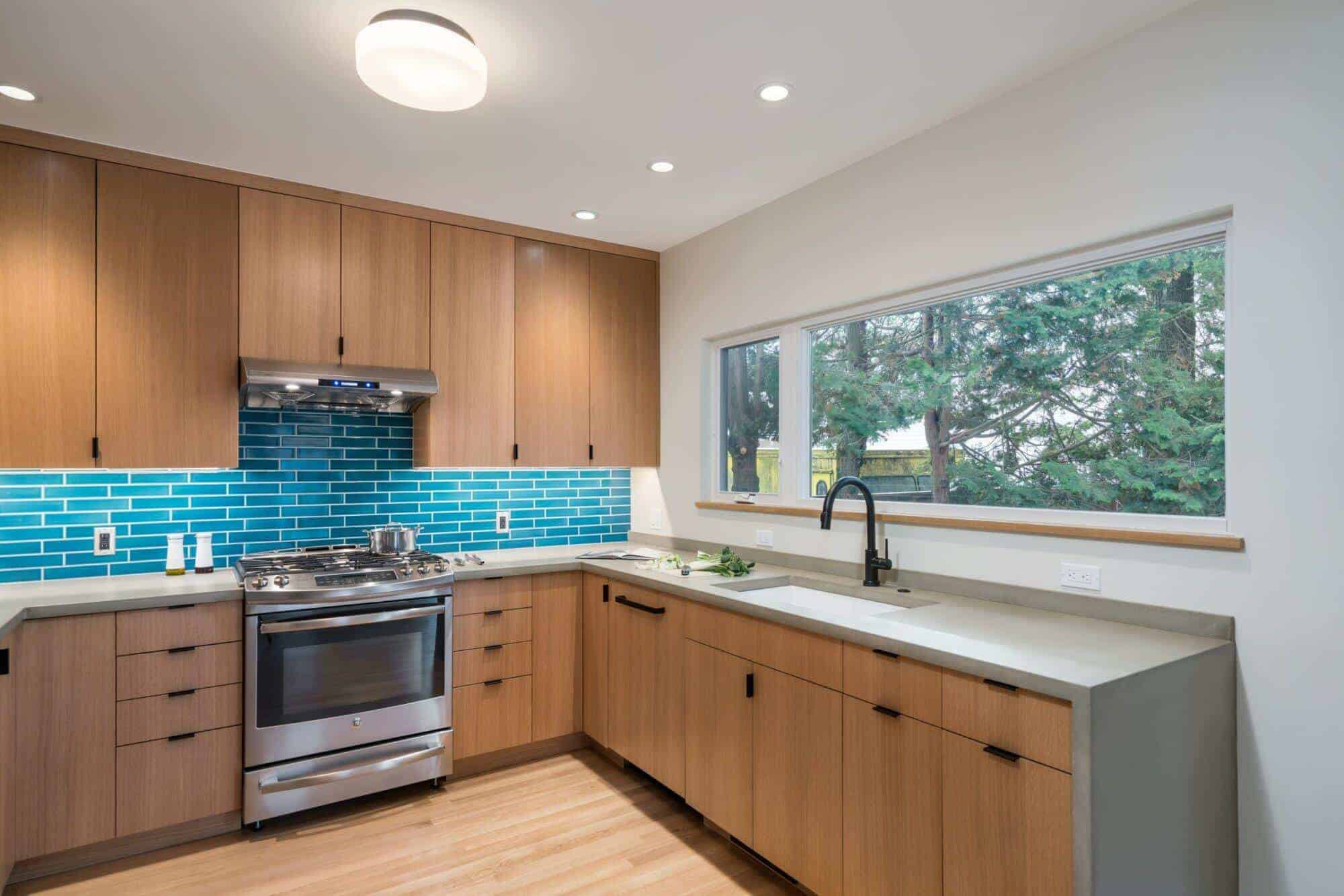 Modern kitchen with light wood cabinets, stainless steel appliances, blue tile backsplash, and large window overlooking trees. Modern kitchen with light wood cabinets, stainless steel appliances, blue tile backsplash, and large window overlooking trees.