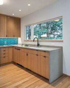 Modern kitchen with wood cabinetry, concrete countertops, a blue tile backsplash, and a large window overlooking trees. Pendant lighting and hardwood floors complete the space.