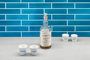 A bottle of The Balvenie DoubleWood 12-Year-Old Single Malt Scotch Whisky on a countertop with four small cups against a blue tiled backsplash.