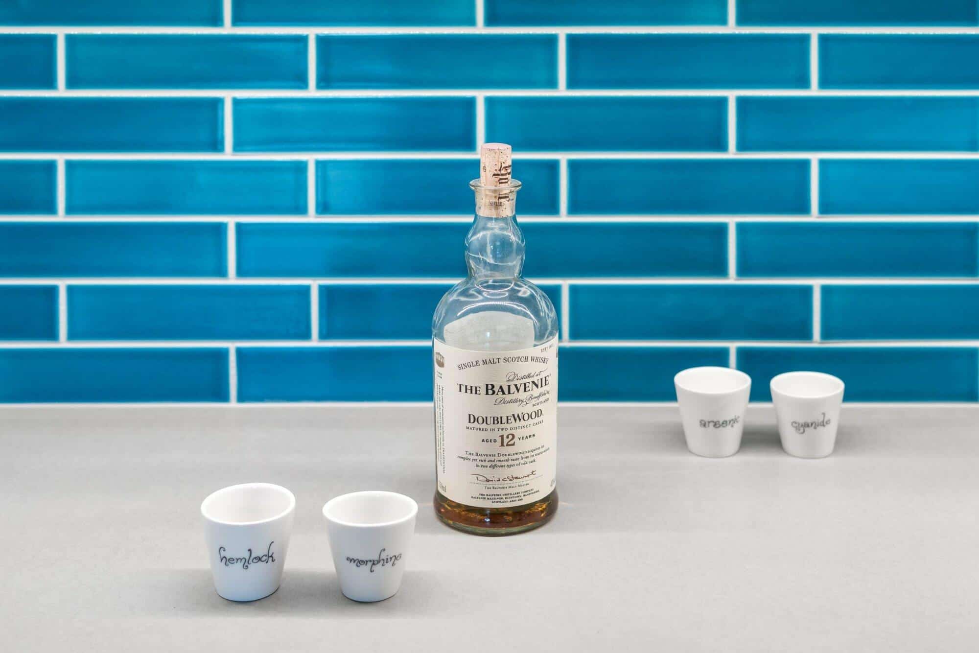 A bottle of The Balvenie DoubleWood 12-Year-Old Single Malt Scotch Whisky on a countertop with four small cups against a blue tiled backsplash. A bottle of The Balvenie DoubleWood 12-Year-Old Single Malt Scotch Whisky on a countertop with four small cups against a blue tiled backsplash.