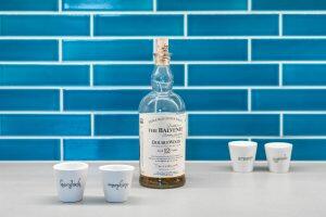 A bottle of The Balvenie DoubleWood 12-year-old Scotch whisky is on a countertop with four white shot glasses labeled 