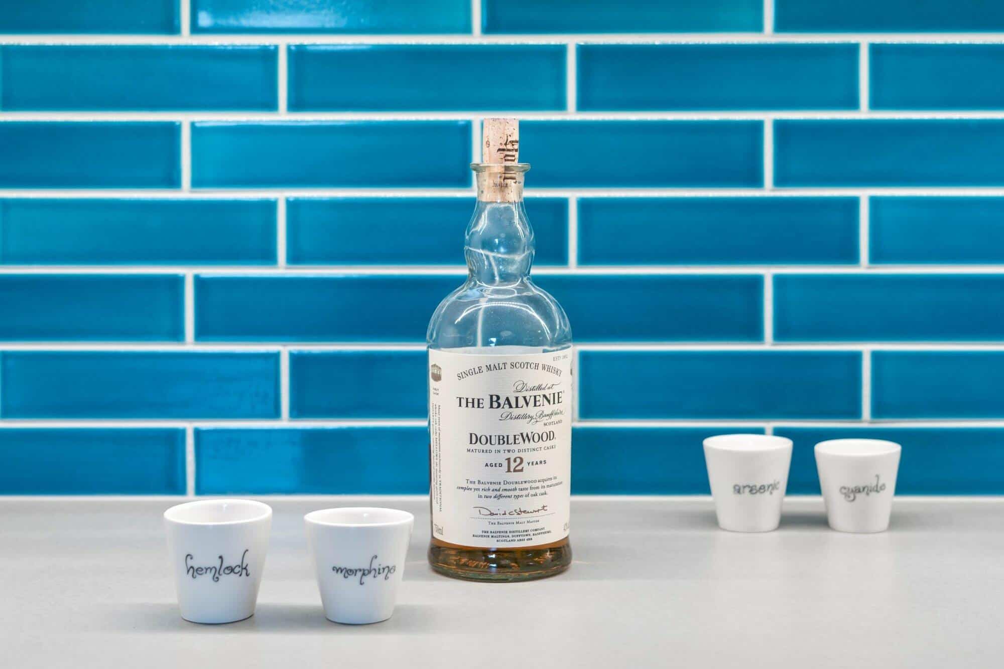 A bottle of The Balvenie DoubleWood 12-year-old Scotch whisky is on a countertop with four white shot glasses labeled A bottle of The Balvenie DoubleWood 12-year-old Scotch whisky is on a countertop with four white shot glasses labeled