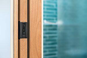 Close-up of a sliding wooden door with a recessed black handle on the left, and a blurry view of a tiled wall in the background on the right.