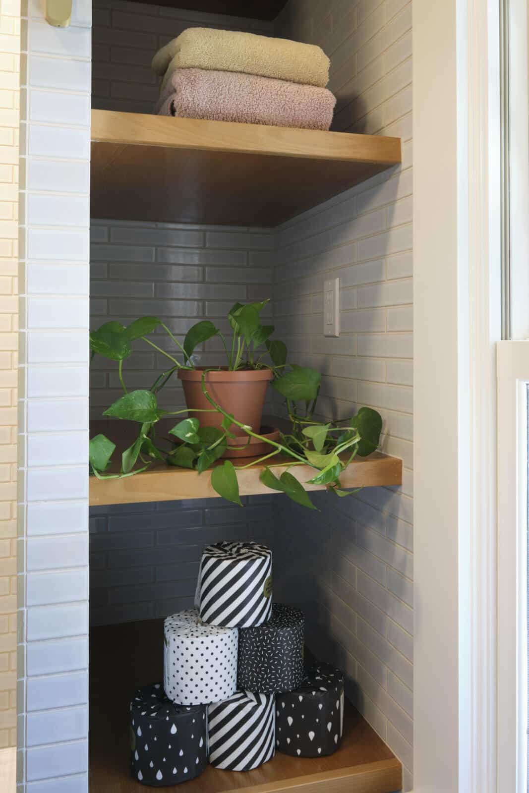 Wooden shelves in a closet hold folded towels, a potted plant with trailing vines, and a stack of patterned toilet paper rolls. The walls feature white tiles and there is a nearby window.