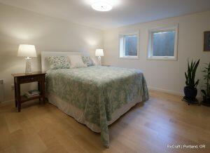 A well-lit bedroom with a double bed, two bedside tables with lamps, green bedding, wooden floor, two small windows, and a potted plant. Text in the corner reads 