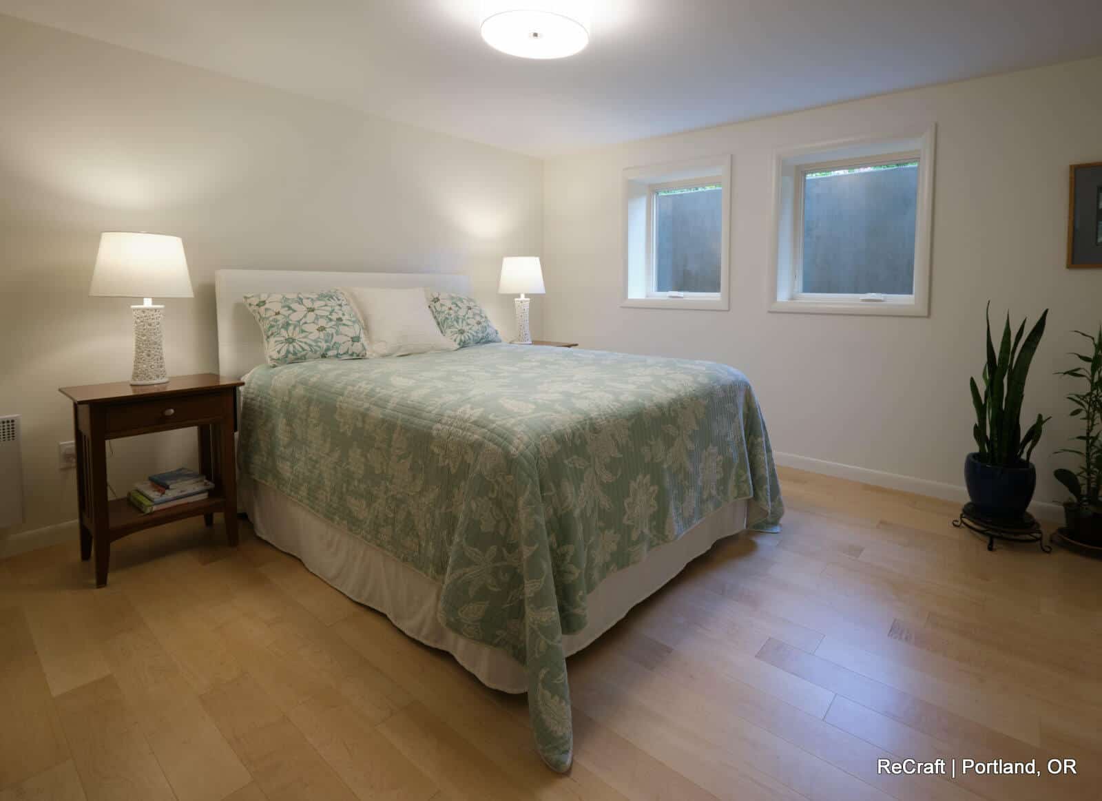Cozy bedroom featuring a queen-sized bed with light green bedding, two bedside lamps, large windows for natural light, and a potted plant, showcasing ReCraft Home Remodeling's basement transformation in Portland.