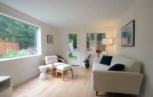 Bright, minimalist living room with a white sofa, wooden chair, small side table, and large window offering a view of a fenced yard with greenery.