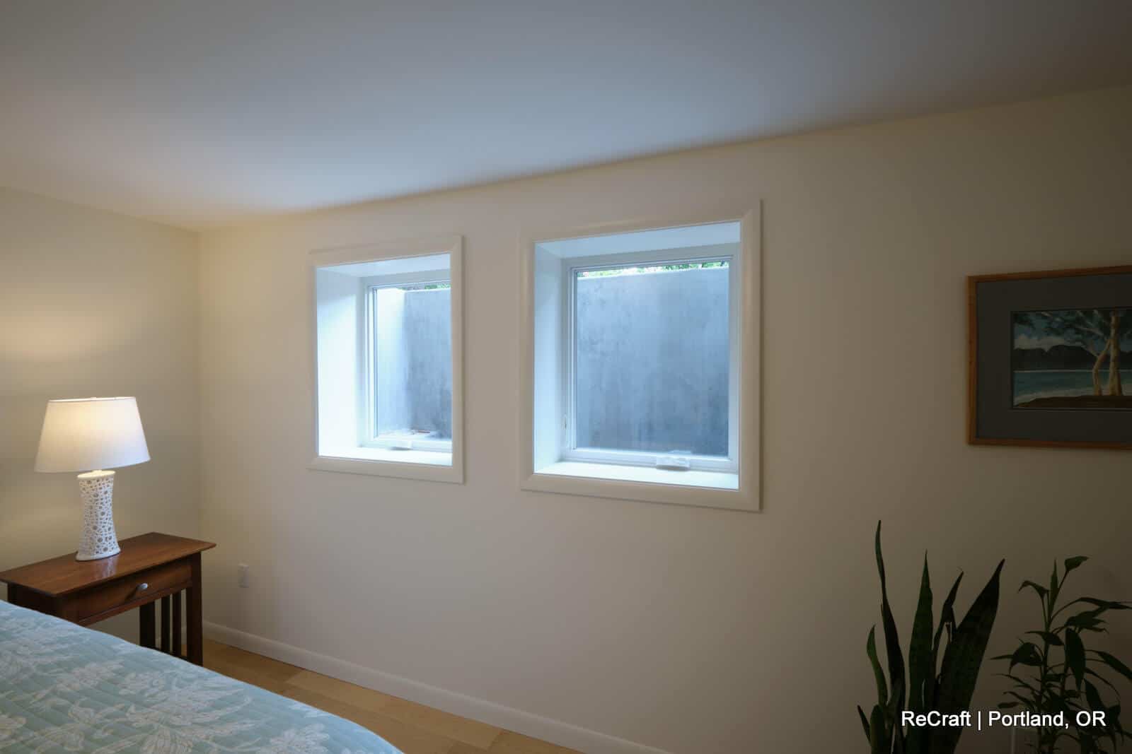 Brightly lit basement room with two windows, modern decor, and a bedside table featuring a lamp, showcasing ReCraft Home Remodeling's basement transformation in Portland.