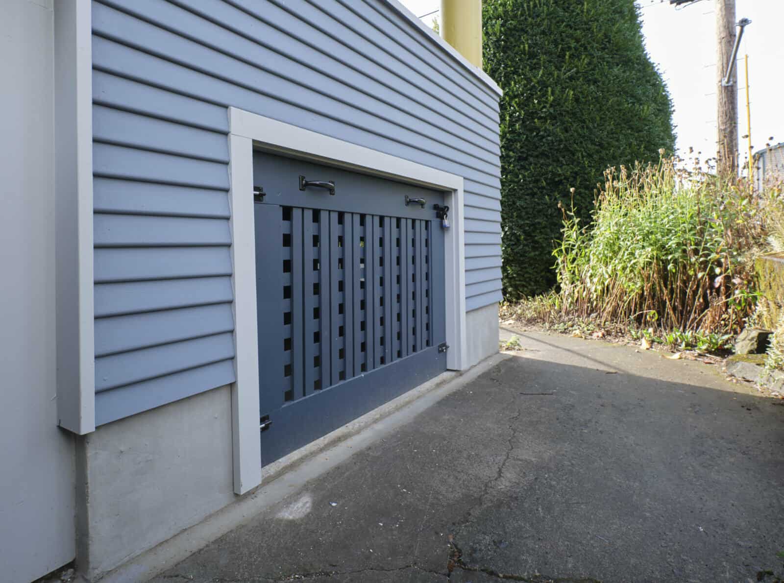 A blue and white horizontal-slatted foundation vent located on the side of a building with bushes and electrical poles in the background.