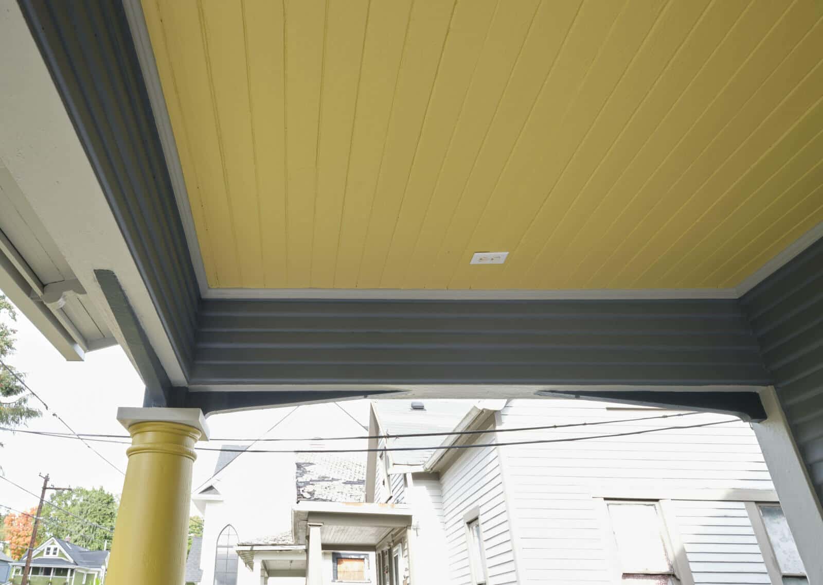 A view of a porch ceiling painted yellow, with the supporting structures painted gray and yellow. An adjacent house and a power line are visible in the background.