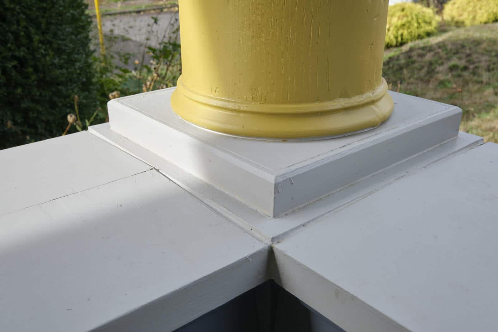 Close-up view of the base of a yellow-painted column set in a white rectangular support structure, situated outdoors with greenery in the background.