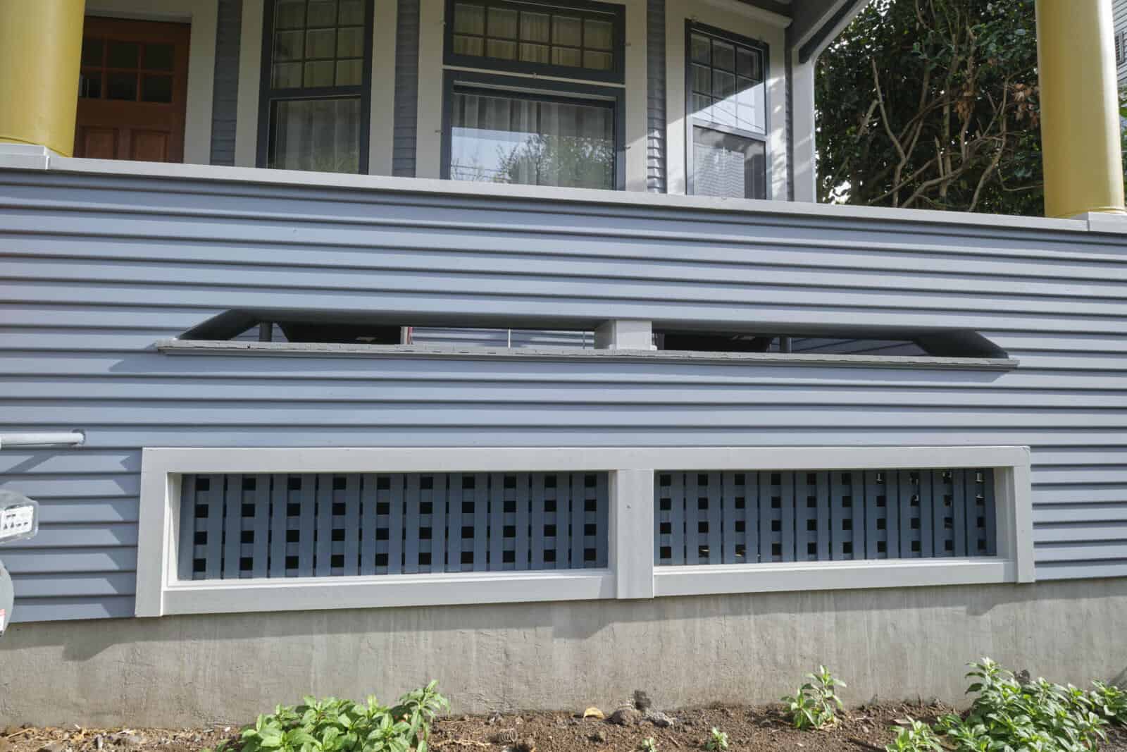 A grey house exterior with horizontal siding features a rectangular vent above two lattice panels. A small garden bed with green plants is in the foreground.