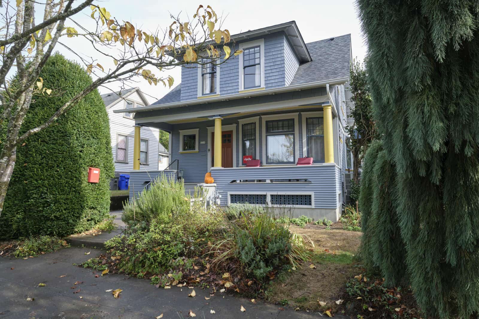 A two-story house with light blue exterior, yellow columns, and a front porch with chairs. It is surrounded by shrubs, trees, and a small garden.