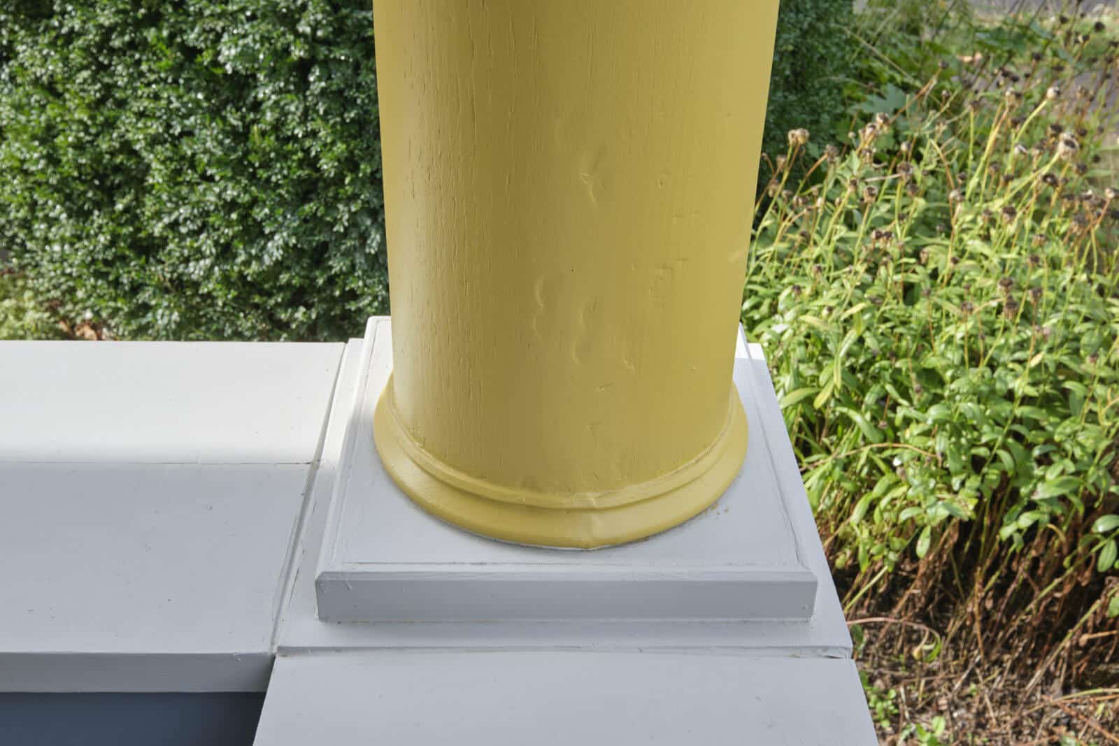 Close-up of the base of a yellow column on a porch with surrounding greenery in the background. The column is secured on a white pedestal.