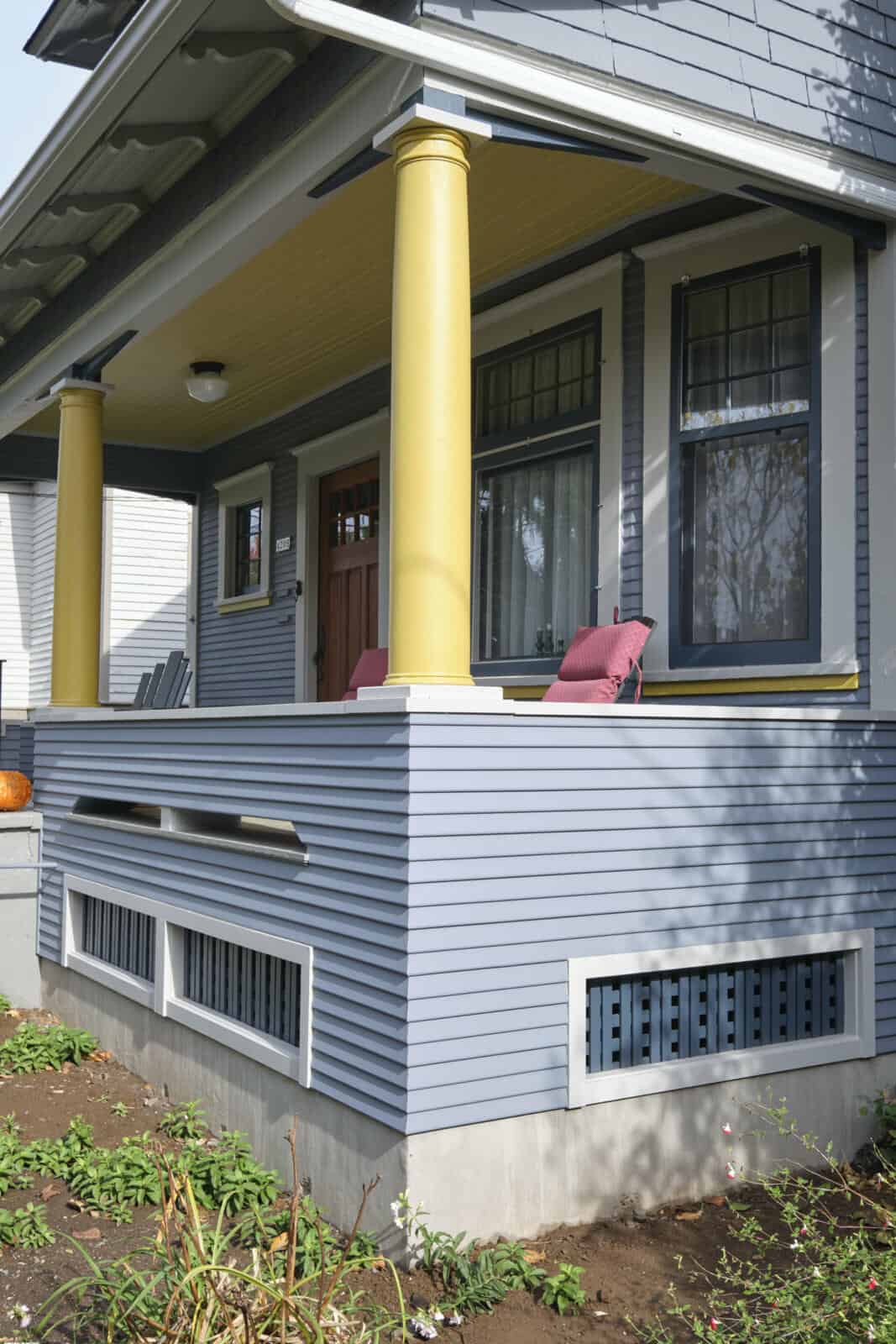 A grey and blue house with a porch featuring two yellow pillars. The porch has two pink cushioned chairs and there is a small green garden in the foreground.