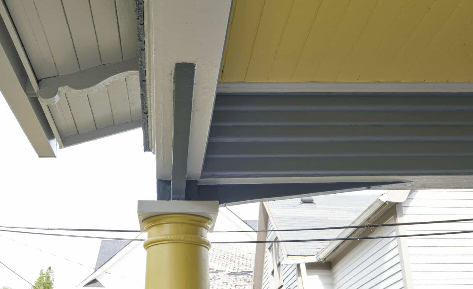 A close-up view of a residential exterior corner showing the intersection of a yellow column, grey support beams, and a yellow ceiling with another house visible in the background.