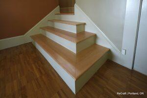 A set of light wooden stairs with white risers against a brown and white wall in a well-lit interior space. The text 