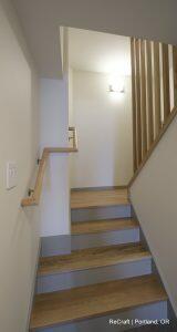 A modern indoor staircase with wooden steps, light-colored walls, and a handrail on the left side. A light fixture is mounted near the top. 