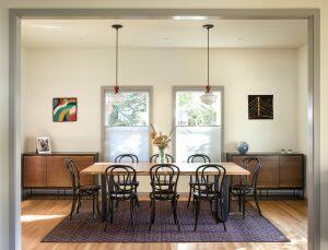 A dining room with a wooden table, eight black chairs, two pendant lights, two cabinets, three artworks on the wall, and a central floral arrangement.