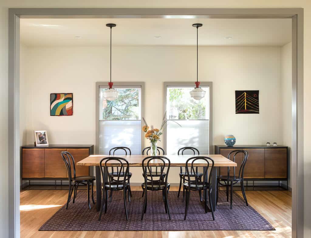 A dining room with a wooden table, eight black chairs, two pendant lights, two cabinets, three artworks on the wall, and a central floral arrangement. A dining room with a wooden table, eight black chairs, two pendant lights, two cabinets, three artworks on the wall, and a central floral arrangement.