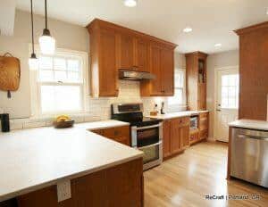 A modern kitchen with wooden cabinets, stainless steel appliances, white countertops, and a white subway tile backsplash. The room has hardwood floors and a back door with a window.