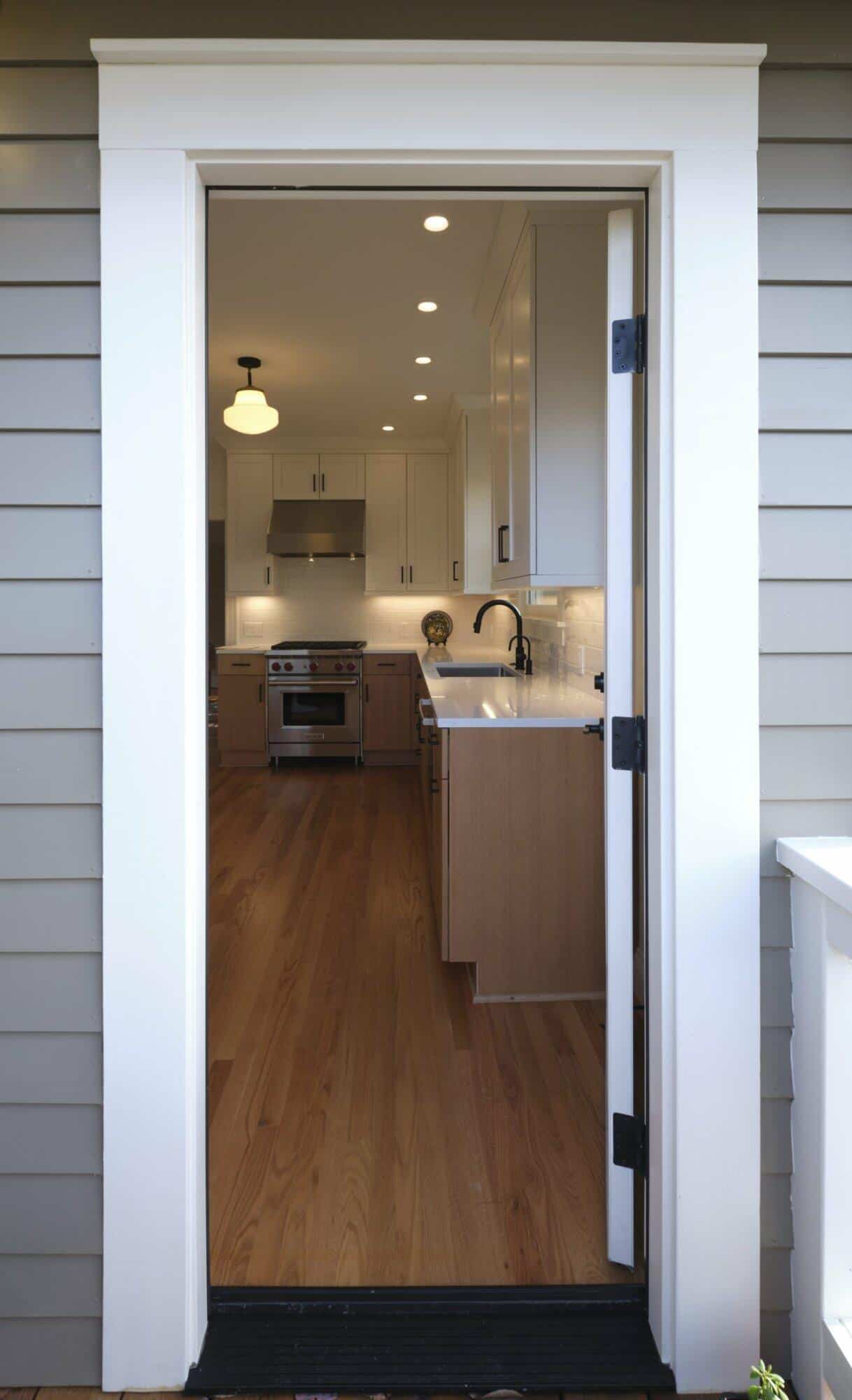 View through an open door into a modern kitchen with wooden floors, white cabinets, stainless steel appliances, and black faucets. View through an open door into a modern kitchen with wooden floors, white cabinets, stainless steel appliances, and black faucets.