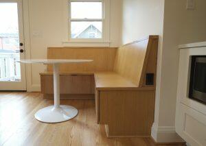 A built-in wooden bench with storage drawers underneath is paired with a white round table in a cozy dining nook near a window and door. The space features light wood flooring and white walls.