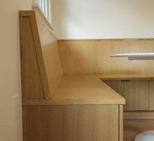 A wooden corner bench with a backrest and no cushions is built into a wall. There is a power outlet on the wall next to the bench. A white table extends from the right side of the image.