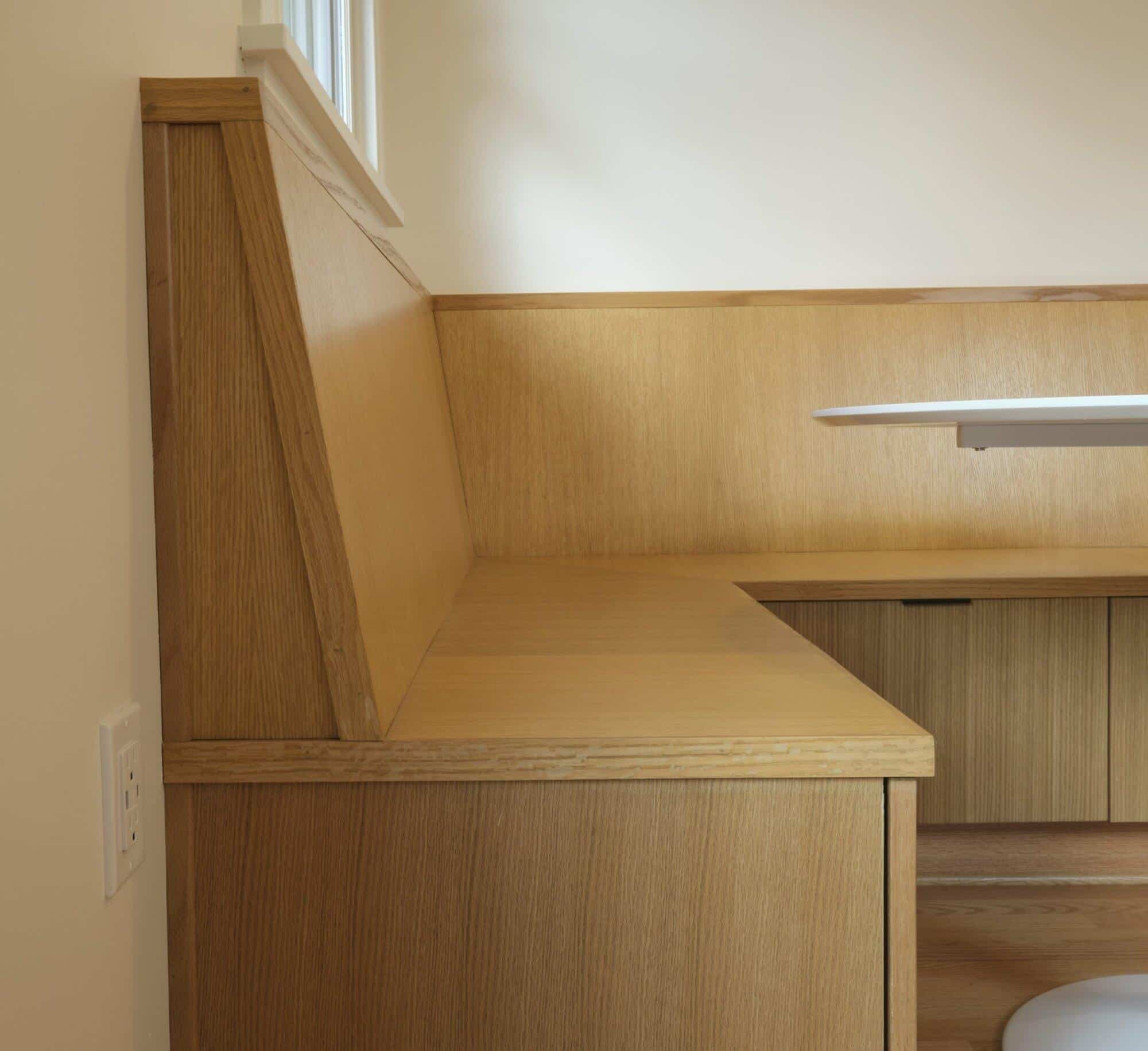 A wooden corner bench with a backrest and no cushions is built into a wall. There is a power outlet on the wall next to the bench. A white table extends from the right side of the image. A wooden corner bench with a backrest and no cushions is built into a wall. There is a power outlet on the wall next to the bench. A white table extends from the right side of the image.