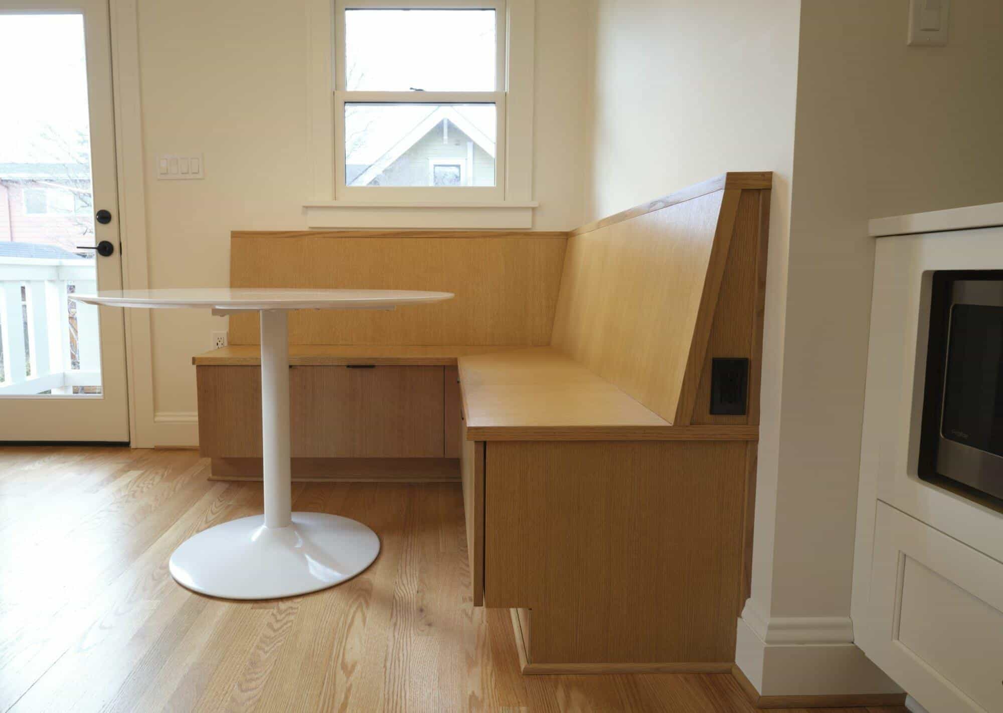 A built-in wooden bench with storage drawers underneath is paired with a white round table in a cozy dining nook near a window and door. The space features light wood flooring and white walls. A built-in wooden bench with storage drawers underneath is paired with a white round table in a cozy dining nook near a window and door. The space features light wood flooring and white walls.