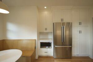Modern kitchen with white cabinets, stainless steel refrigerator, built-in microwave, and wooden seating area with a round white table. Light wood flooring and recessed lighting are visible.