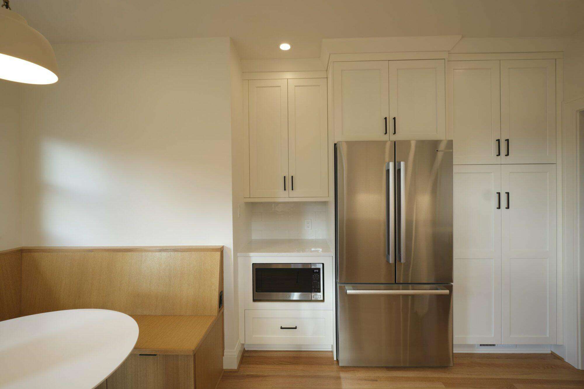 Modern kitchen with white cabinets, stainless steel refrigerator, built-in microwave, and wooden seating area with a round white table. Light wood flooring and recessed lighting are visible. Modern kitchen with white cabinets, stainless steel refrigerator, built-in microwave, and wooden seating area with a round white table. Light wood flooring and recessed lighting are visible.