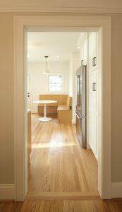 View through a doorway into a kitchen with wooden floors, white cabinets, a stainless steel refrigerator, and a built-in dining nook with a round table and window.
