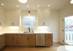 Modern kitchen with wood and white cabinetry, stainless steel dishwasher, black faucet, and recessed lighting. A window and glass door provide natural light.