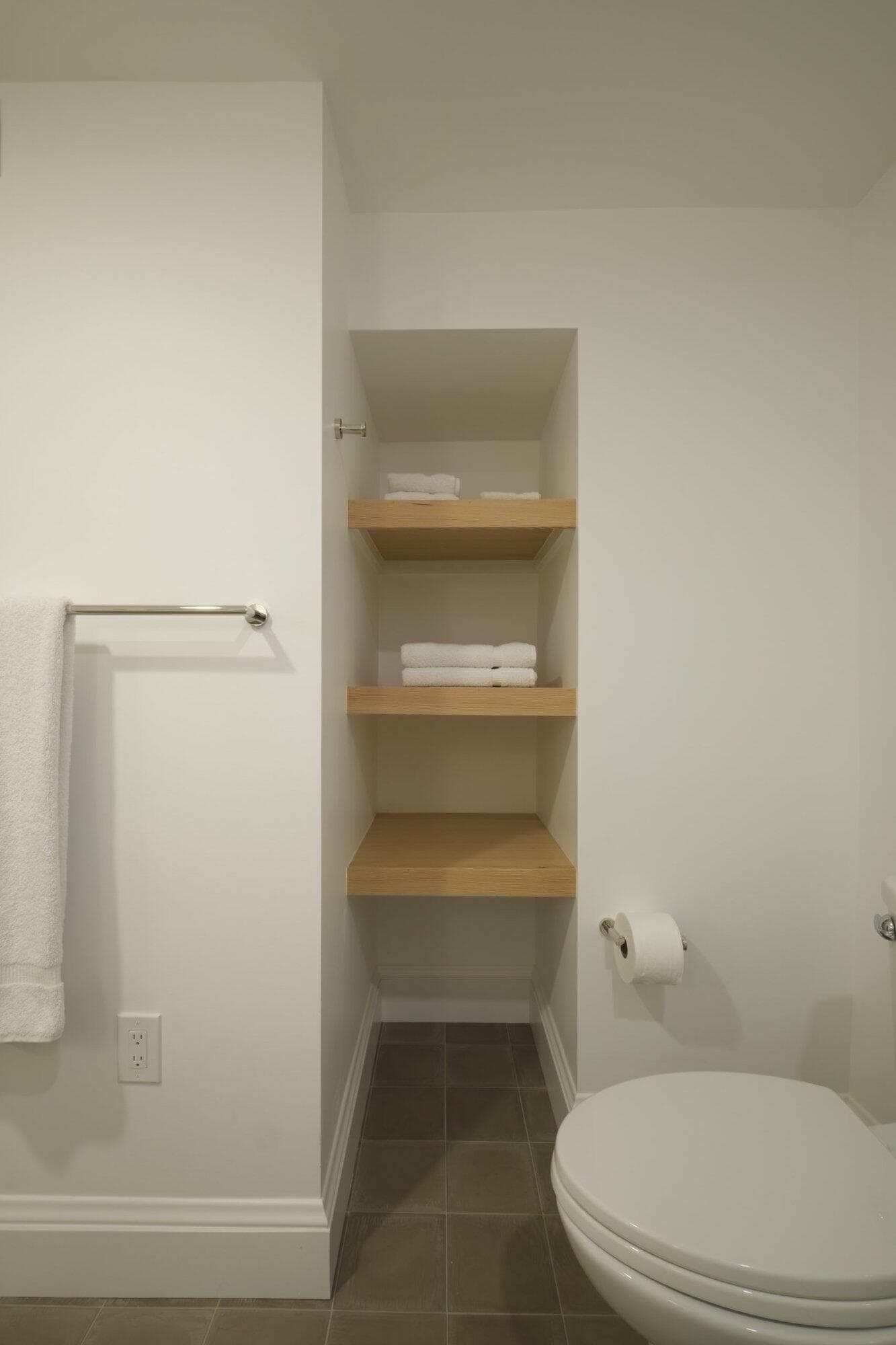 Bathroom interior with white walls, a towel on a towel rack, three wooden shelves holding folded white towels, and a toilet next to a roll of toilet paper.