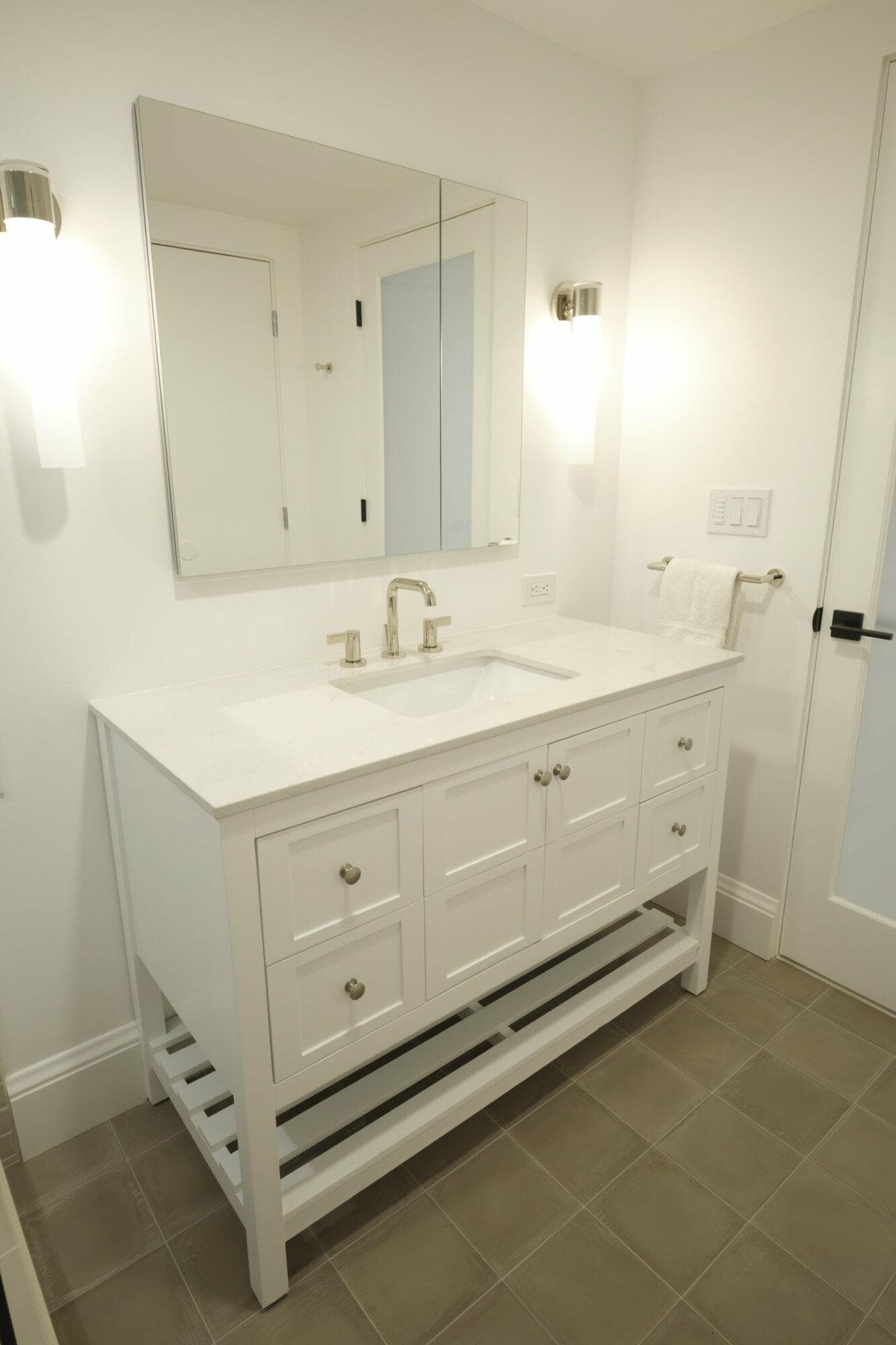 A modern bathroom with a white vanity, countertop, and sink, featuring a large mirror, two wall-mounted lights, and a towel ring beside the sink on a tiled floor.