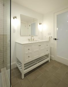 A bathroom with a white sink cabinet, mounted mirror, towel rack, and glass shower door. The room has beige floor tiles and minimalistic decor.